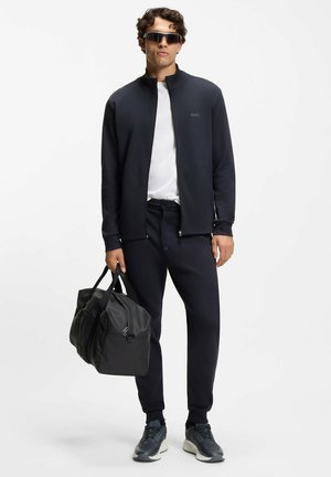 Giacca navy con zip sopra una maglietta bianca, abbinata a pantaloni jogger navy. Borsone nero in mano, scarpe da ginnastica grigie con texture.