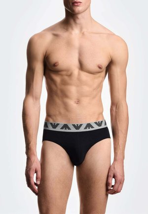 Modello maschile senza maglietta che indossa slip neri con una fascia grigia in vita con un motivo triangolare ripetuto, in piedi davanti a uno sfondo semplice.