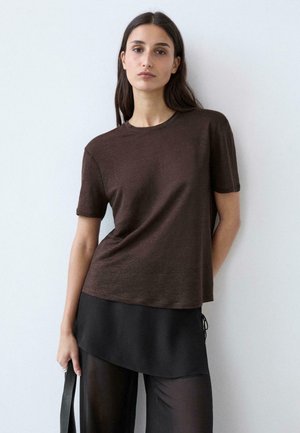 Femme aux longs cheveux noirs portant un haut marron à manches courtes avec une jupe noire superposée et un pantalon noir transparent, tenant une sangle noire.