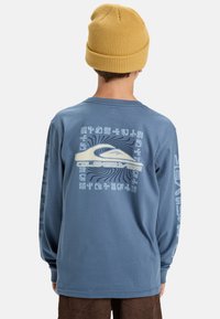 Långärmad blå t-shirt med en grafisk design på ryggen som visar en våg och text. Bärs med en gul stickad mössa.