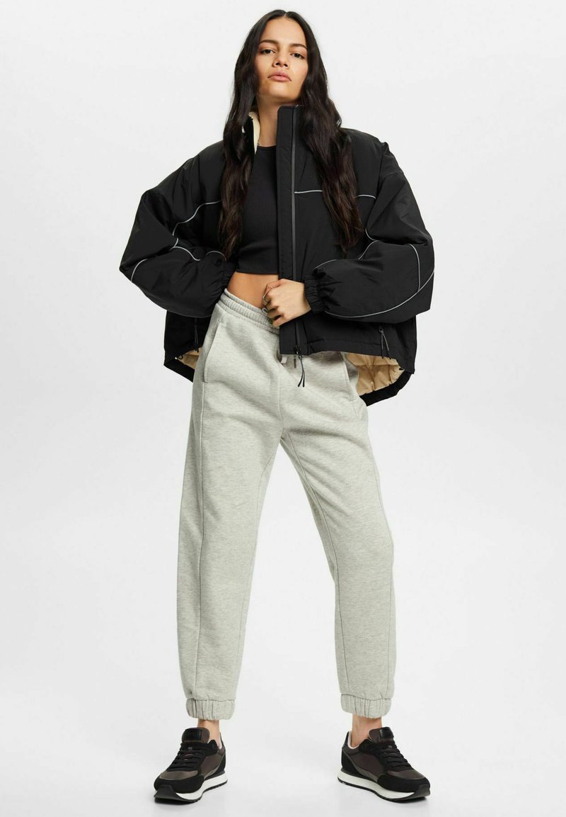 Giacca imbottita nera con cuciture bianche, abbinata a pantaloni della tuta grigi chiari. Sneakers in toni scuri con suole bianche completano il look.