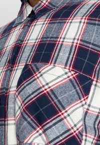Camicia a quadri nei colori blu navy, bianco e rosso. Include un colletto con bottoni, un motivo a scacchi e un dettaglio con taschino sul petto. Tessuto morbido.