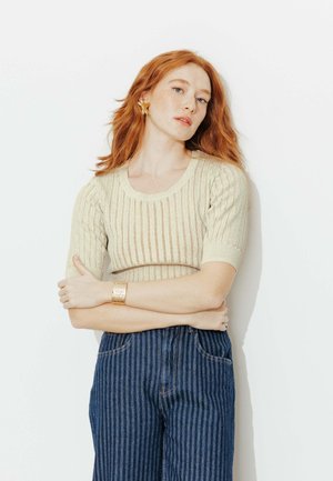 Femme aux cheveux roux avec de légères taches de rousseur, portant un haut en maille crème et un pantalon bleu à fines rayures, debout contre un fond blanc les bras croisés.