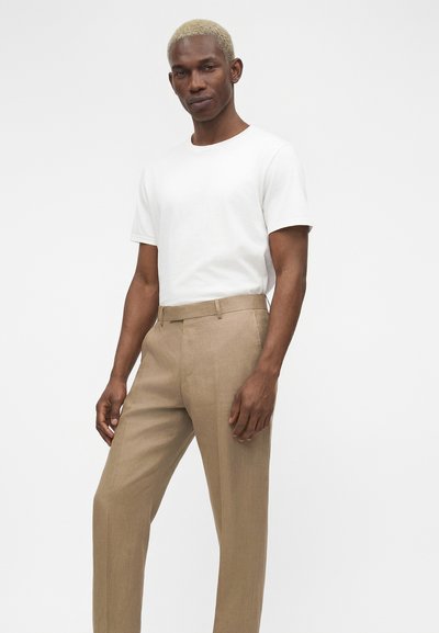 Άνδρας με κοντά ξανθά μαλλιά που φοράει λευκό κοντομάνικο μπλουζάκι και μπεζ ψηλόμεσο παντελόνι στέκεται μπροστά σε απλό φόντο.