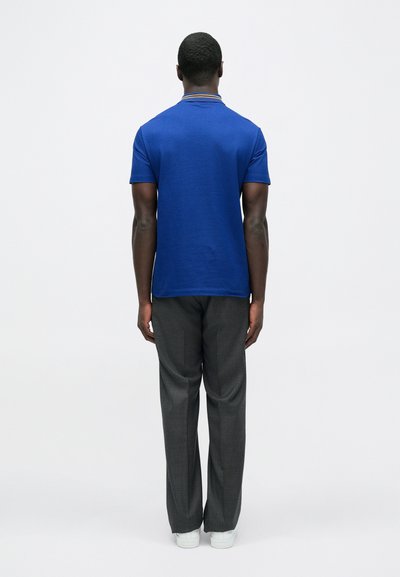 Mėlyna polo marškinėliai su kontrastine apykakle, derinami su pilkais languotais kelnėmis ir baltais sportbačiais, žiūrint iš nugaros.