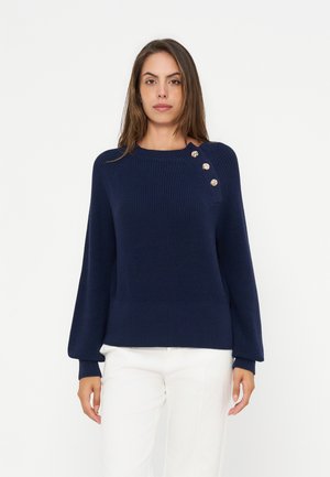 Ung kvinna som bär en marinblå stickad tröja med dekorativa knappar på axeln och vita byxor, stående mot en enfärgad bakgrund.