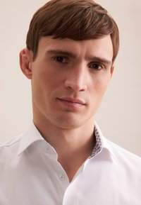 Biała koszula zapinana na guziki z wzorzystym kołnierzykiem, gładką fakturą i usztywnionym krojem. Zbliżenie na twarz męskiego modela, neutralne tło.