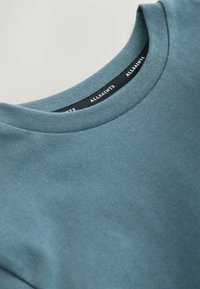 Vaaleansininen puuvillainen pyöreäkauluksinen t-paita, jossa on sileä pinta. Kauluksen sisäpuolella on musta etiketti, jossa lukee "ALLSAINTS".