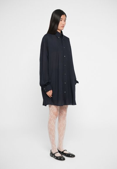 Vestido camiseiro azul-marinho com botões, mangas longas, tecido leve e corte relaxado, combinado com meias-calças nudes estampadas e sapatilhas pretas.