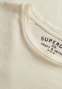 Tricou din bumbac de culoare crem cu decolteu rotund. Prezintă o etichetă cu inscripția "SUPERDRY CO. CRAFT & DETAIL" în text negru. Textură moale.
