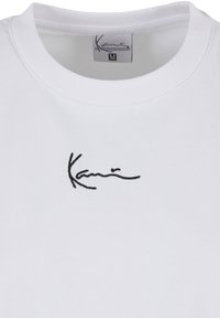 Tricou alb cu guler rotund, cu broderie neagră în stil cursiv „Karni” centrată sub linia gâtului, etichetă de mărime medie în interiorul gulerului.