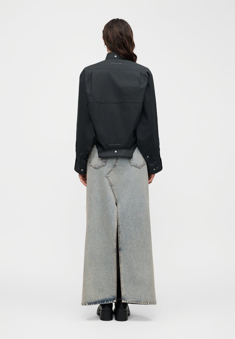 Personne portant une veste noire boutonnée et une longue jupe en denim clair avec une fente à l'arrière, ainsi que des bottes noires, debout de dos.