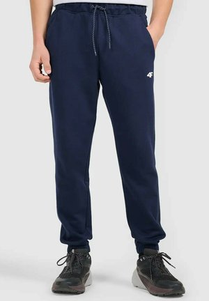 Mężczyzna ubrany w granatowe dresowe spodnie z sznurkiem w pasie oraz czarne buty sportowe, stojący na jednolitym tle.