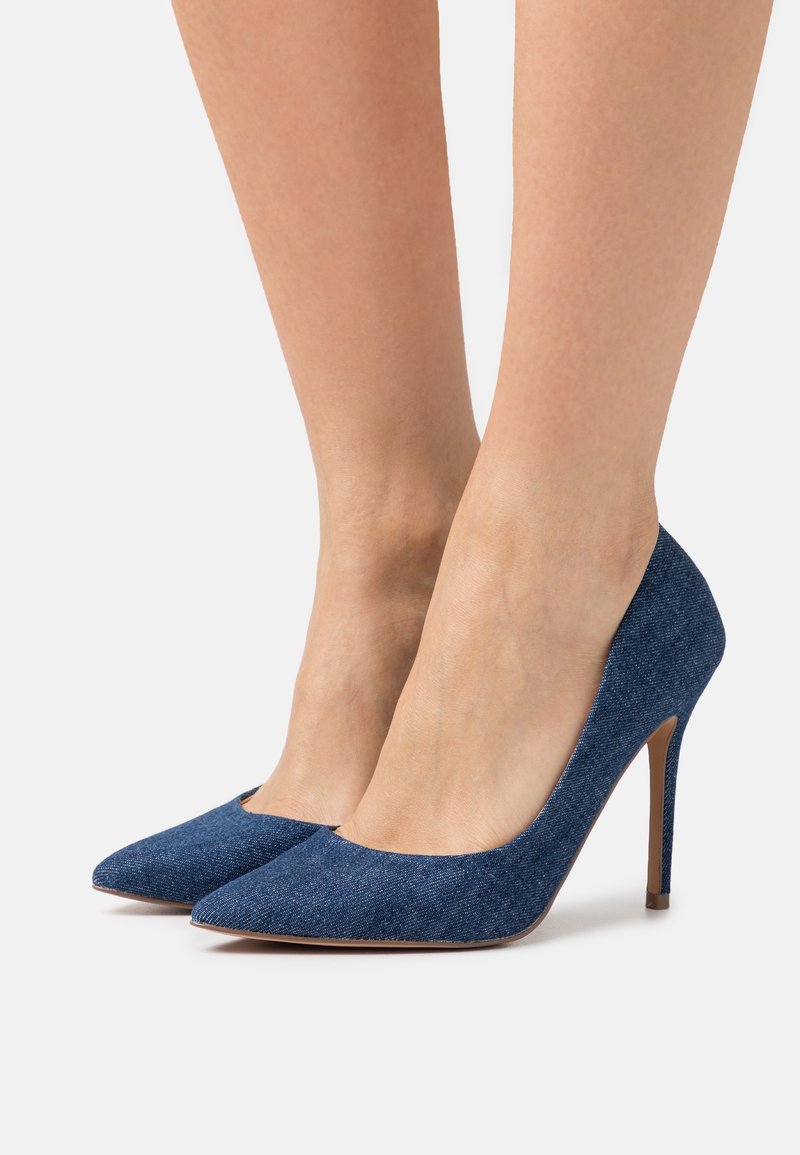 Femme portant des escarpins à talons hauts en jean bleu à bout pointu, debout sur fond blanc. Seules les jambes inférieures et les chaussures sont visibles.