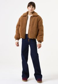 Chaqueta marrón, esponjosa y de gran tamaño con un cuello, cierre de cremallera en la parte delantera y puños acanalados, llevada sobre un suéter crema y unos pantalones de campana azul oscuro.