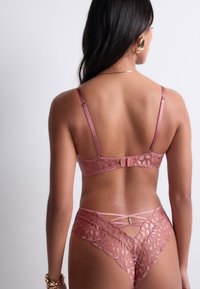 Femme vue de dos portant un ensemble de lingerie en dentelle rose avec des accents dorés, un collier doré, des boucles d'oreilles dorées et des bracelets dorés.