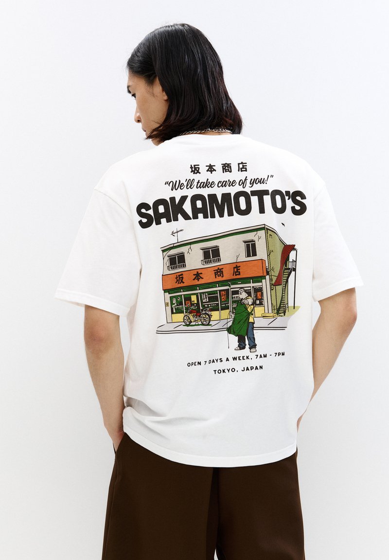 Bílý bavlněný tričko s grafickým potiskem obchodu s zelenými akcenty, textem "SAKAMOTO'S" a postavou držící tašku.