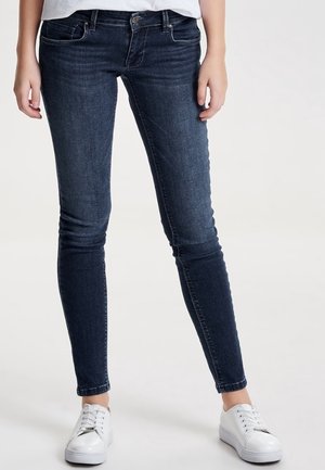 Personne portant un jean skinny bleu foncé et des baskets blanches, debout devant un fond clair uni, les mains détendues le long du corps.