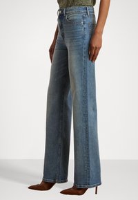 Plačios mėlynos džinsinės kelnės, pasižyminčios aukštu liemeniu, subtiliu blukimu ir klasikiniais penkių kišenių modeliais, derinamos su rudais smailiais bateliais.