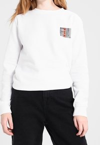Vit sweatshirt med rund halsringning och långa ärmar. Har en liten svart och röd grafisk text "OFF THE WALL" på vänster bröst.