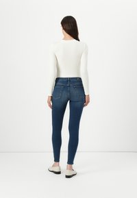 Un jean skinny bleu foncé à taille haute, avec deux poches arrière et une texture lisse, associé à un haut ajusté à manches longues blanc.