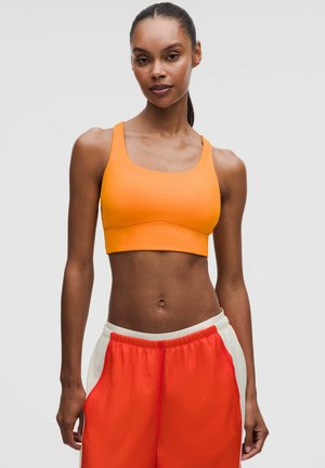 Kvinde iført en orange sports-bh og røde atletikshorts med hvide sidepaneler, stående mod en ensfarvet lys baggrund.