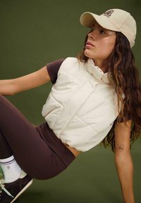 Puffig beskuren väst i off-white tyg med hög krage, kombinerad med en chokladbrun åtsittande topp och matchande leggings, mot en grön bakgrund.