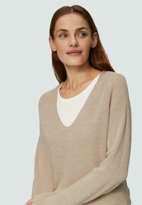 Beige, gestrickter Pullover mit tiefem V-Ausschnitt und langen Ärmeln; weiche Textur und Rippendetails an den Ärmeln, über einem weißen Hemd getragen.