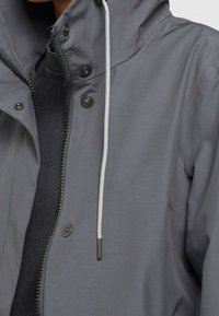 Detail zblízka na osobu v šedé bundě s černými knoflíky, zipem a bílým šňůrkovým stahováním, pod kterou má černé tričko.