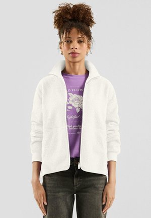 Frau mit lockigem Haar trägt eine weiße Fleecejacke mit Reißverschluss über einem lila T-Shirt mit Aufdruck und dunklen Jeans, steht vor einem schlichten Hintergrund.