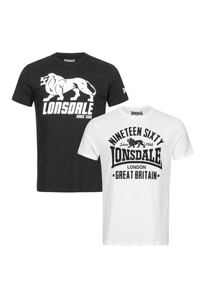 Kopasz férfi nyak- és kar tetoválásokkal, fekete Lonsdale pólóban, sötét farmerral és fehér tornacipővel, szürke háttér előtt áll.