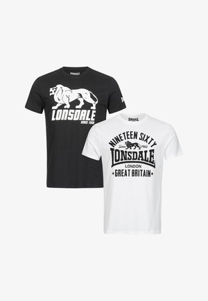 Kopasz férfi nyak- és kar tetoválásokkal, fekete Lonsdale pólóban, sötét farmerral és fehér tornacipővel, szürke háttér előtt áll.