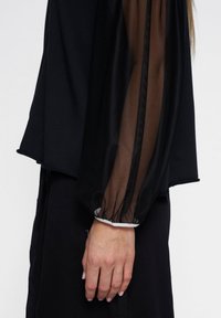 Zwarte top met doorzichtige zwarte mesh mouwen, elastische manchetten en een losse pasvorm. Het materiaal is glad met een contrasterende textuur in de mouwen.