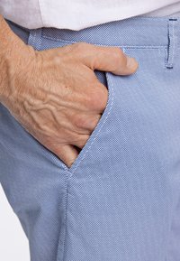 Pantaloni azzurri testurizzati con un tessuto a fantasia, caratterizzati da una tasca laterale e una mano visibile che riposa nella tasca.
