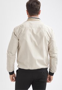 Beige leichte Bomberjacke mit gerippten schwarzen Bündchen und Kragen, mit glatter Textur und elastischem Bund, von hinten gezeigt.