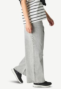 Helle graue weit geschnittene Jeans mit gefleckter Textur, kombiniert mit einem schwarz-weißen gestreiften Oberteil und schwarzen Sportschuhen.