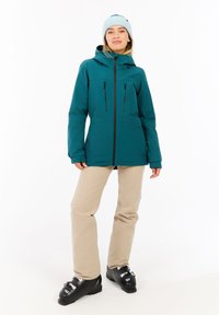 Türkisfarbener wasserdichter Jacke mit Kapuze, Frontreißverschluss und Reißverschlusstaschen, kombiniert mit beigen Schneehosen und schwarzen Skischuhen.