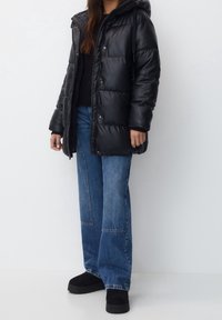 Veste matelassée noire avec capuche, étincelante, boutons-pression et poches latérales, associée à un jean bleu ample et des chaussures à plateforme noires.