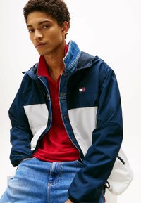 Chaqueta azul marino y blanca con cuello de mezclilla, que presenta un parche con un logo. Lleva puesta una camiseta polo roja y se combina con jeans azules. Textura suave.