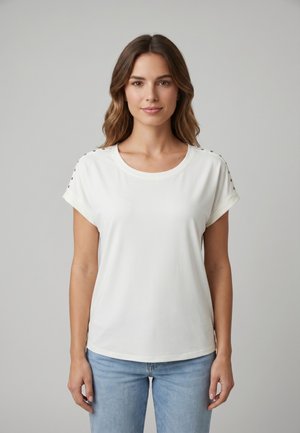 Mujer con cabello castaño ondulado que lleva una camisa blanca de manga corta con botones en los hombros y vaqueros azul claro, de pie frente a un fondo gris.