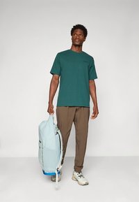 T-shirt verde de mangas curtas com decote redondo, combinada com calças castanhas claras e uma mochila azul clara. Ajuste padrão e textura suave.