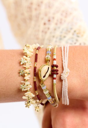 Cinq bracelets au poignet : coquillage, ivoire, et accents ton or, perles rouges et bleues, motifs floraux, et un fond texturé ton pêche.