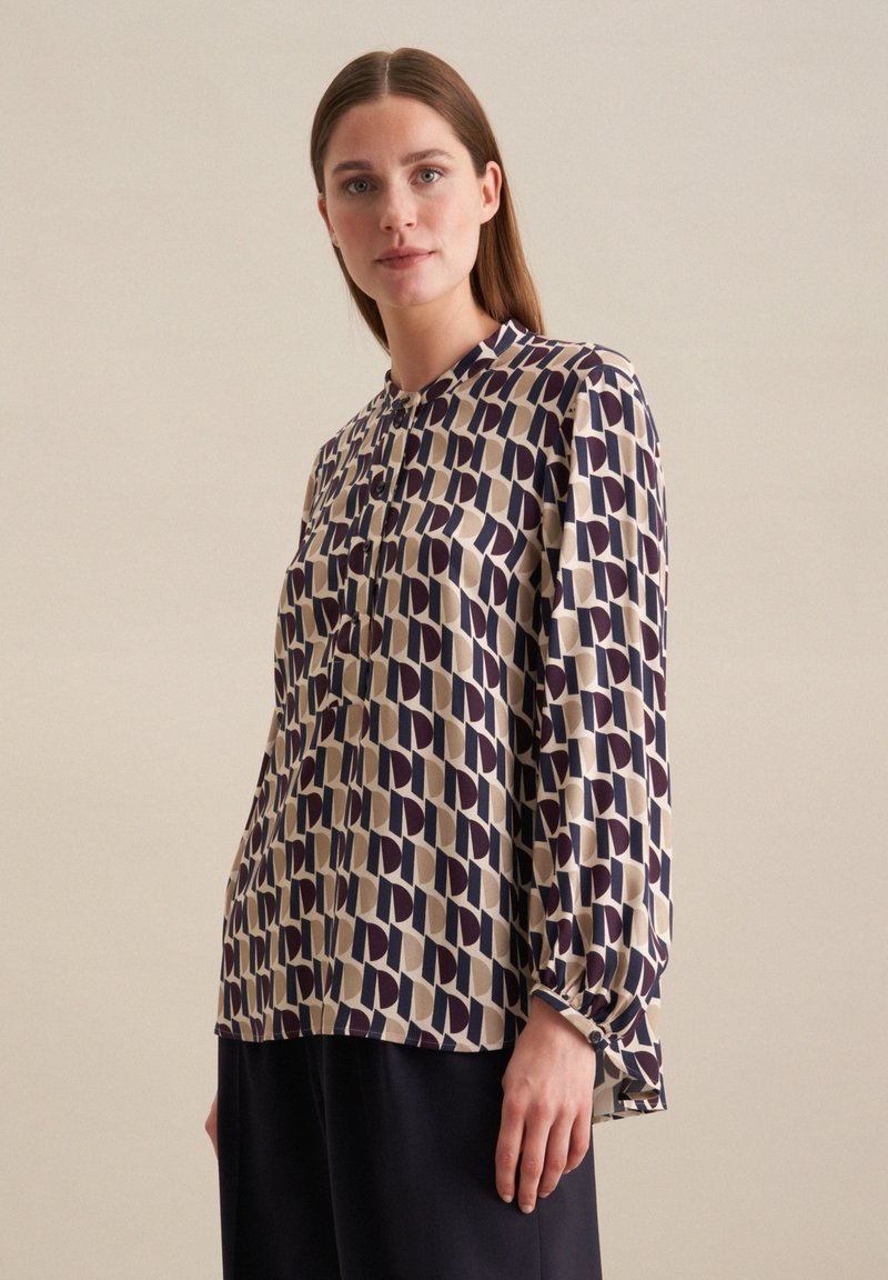 Blouse à manches longues avec un col montant, présentant un motif géométrique en marine et beige. Tissu doux avec des fermetures à boutons aux poignets.