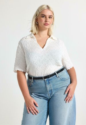 Lyshåret kvinde iført en tekstureret hvid kortærmet skjorte og lyseblå højtaljede jeans med sort bælte, stående mod en ensfarvet baggrund.