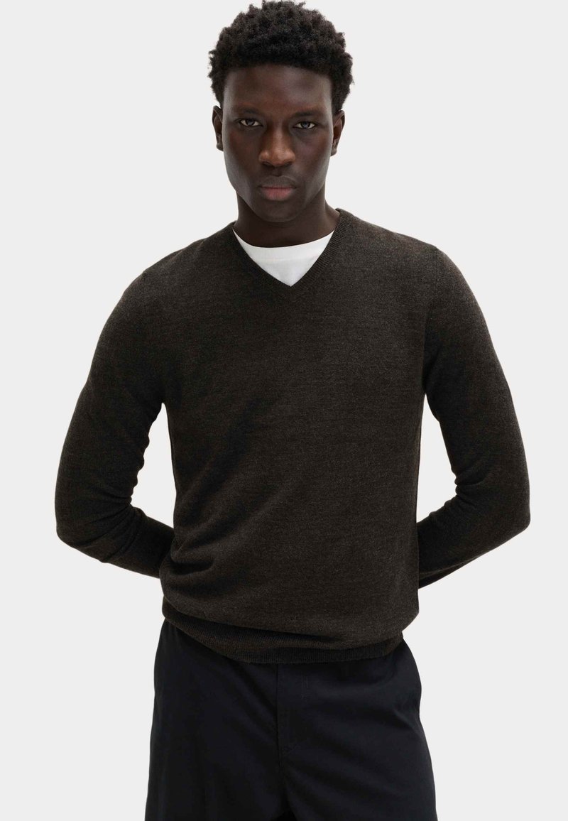 Mężczyzna złożonymi dłońmi, ubrany w ciemnobrązowy sweter z dekoltem w serek na białej koszuli i ciemnych spodniach, na jasnym, jednolitym tle.