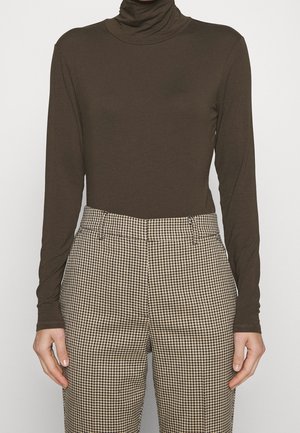 Persoon draagt een bruin turtleneck met lange mouwen en een beige met bruine geruite broek met hoge taille, staat met ontspannen armen langs het lichaam.