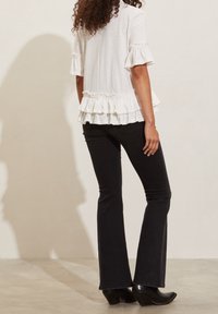 Bílý volánkový top s krátkými rukávy a rozšířeným lemováním, spárovaný s černými rozšířenými džíny a černými koženými botami na podpatku. Hladká textura látky.