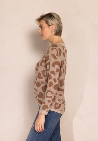 Beiger Pullover mit braunen Leopardenflecken, strukturierter Strick, Rundhalsausschnitt und langen Ärmeln. Kombiniert mit blauen Jeans, im Seitenprofil präsentiert.