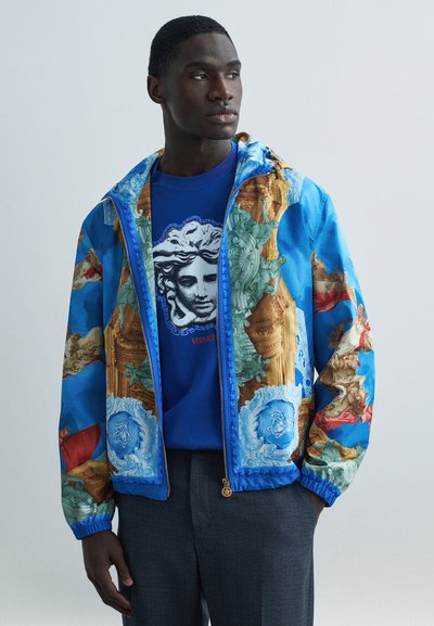 Veste aviateur bleue avec des motifs classiques ornés, comprenant des accents dorés et une fermeture éclair à l'avant. En dessous, un T-shirt bleu avec un imprimé graphique.