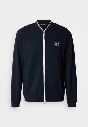 Navy zip-up jacket made of smooth fabric with white zipper, ribbed collar, and EA7 logo on the chest. Side pockets for storage.
